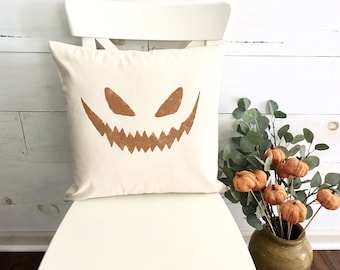 May include: A white pillow with a brown jack-o'-lantern face printed on it. The pillow is sitting on a white chair. There is a vase with orange pumpkins and green leaves in the background.