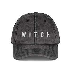May include: A dark gray baseball cap with the word "WITCH" embroidered in white block letters across the front. The cap has a curved brim and a distressed, vintage look.