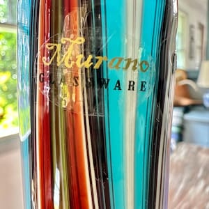 K&ouml;nnte beinhalten: Eine farbenfrohe Murano-Glasvase mit vertikalen Streifen in Rot, Orange, T&uuml;rkis, Schwarz, Wei&szlig; und Gelb. Die Vase hat einen transparenten Aufkleber mit goldfarbener Aufschrift "Murano GLASSWARE". Das Glas ist leicht durchscheinend.