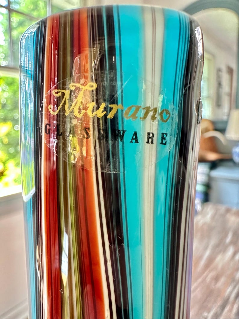 K&ouml;nnte beinhalten: Eine farbenfrohe Murano-Glasvase mit vertikalen Streifen in Rot, Orange, T&uuml;rkis, Schwarz, Wei&szlig; und Gelb. Die Vase hat einen transparenten Aufkleber mit goldfarbener Aufschrift "Murano GLASSWARE". Das Glas ist leicht durchscheinend.