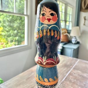 May include: A hand-painted wooden nesting doll with a blue and black color scheme. The doll features a painted face with rosy cheeks and a red mouth. The doll has a layered design with gold and orange accents.
