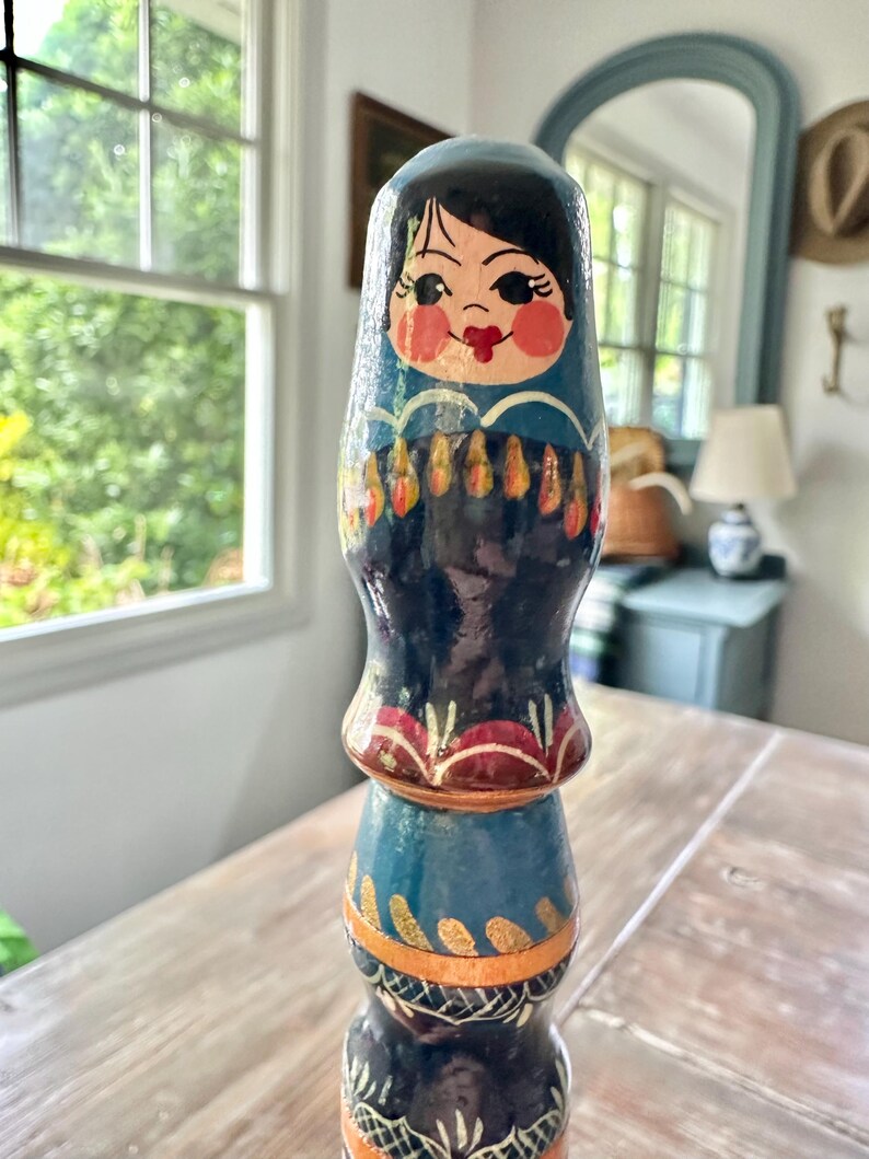 May include: A hand-painted wooden nesting doll with a blue and black color scheme. The doll features a painted face with rosy cheeks and a red mouth. The doll has a layered design with gold and orange accents.