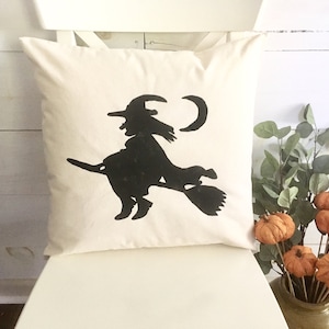 May include: A white pillow with a black silhouette of a witch flying on a broomstick with a crescent moon in the background.