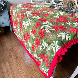 May include: A green tablecloth with a red and white floral pattern, featuring red pom-pom trim. The tablecloth is draped over a white table, creating a festive holiday theme. The pattern includes poinsettias and pinecones.