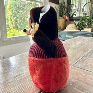 May include: A vintage plush toy in the shape of a cartoon character. The toy has a black and red color scheme, with a red body and black striped top. The toy is positioned on a wooden table.