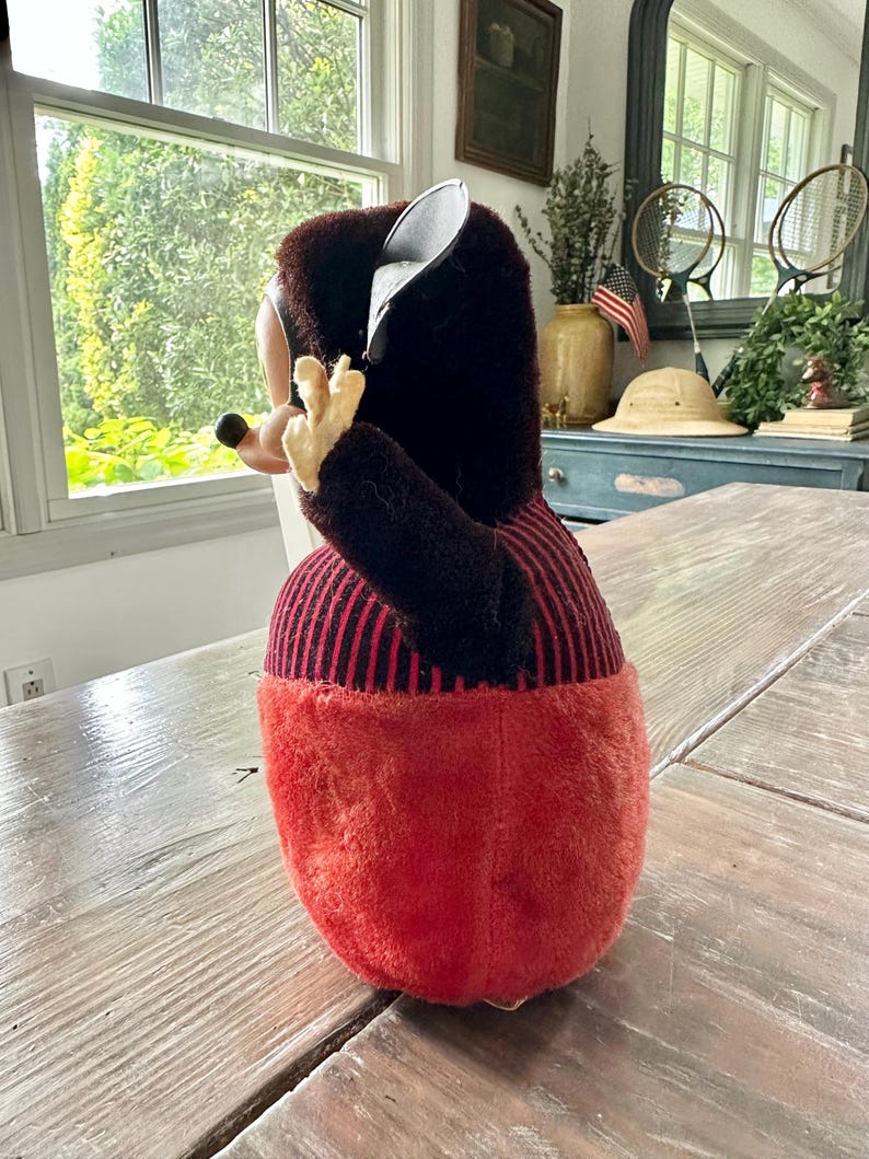 May include: A vintage plush toy in the shape of a cartoon character. The toy has a black and red color scheme, with a red body and black striped top. The toy is positioned on a wooden table.