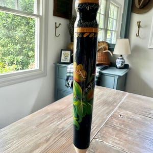 May include: A tall, decorative pencil with a painted floral design. The pencil is primarily black with colorful accents of orange, green, and blue. The pencil's tip is a natural wood color, and the top is painted with red and gold details.