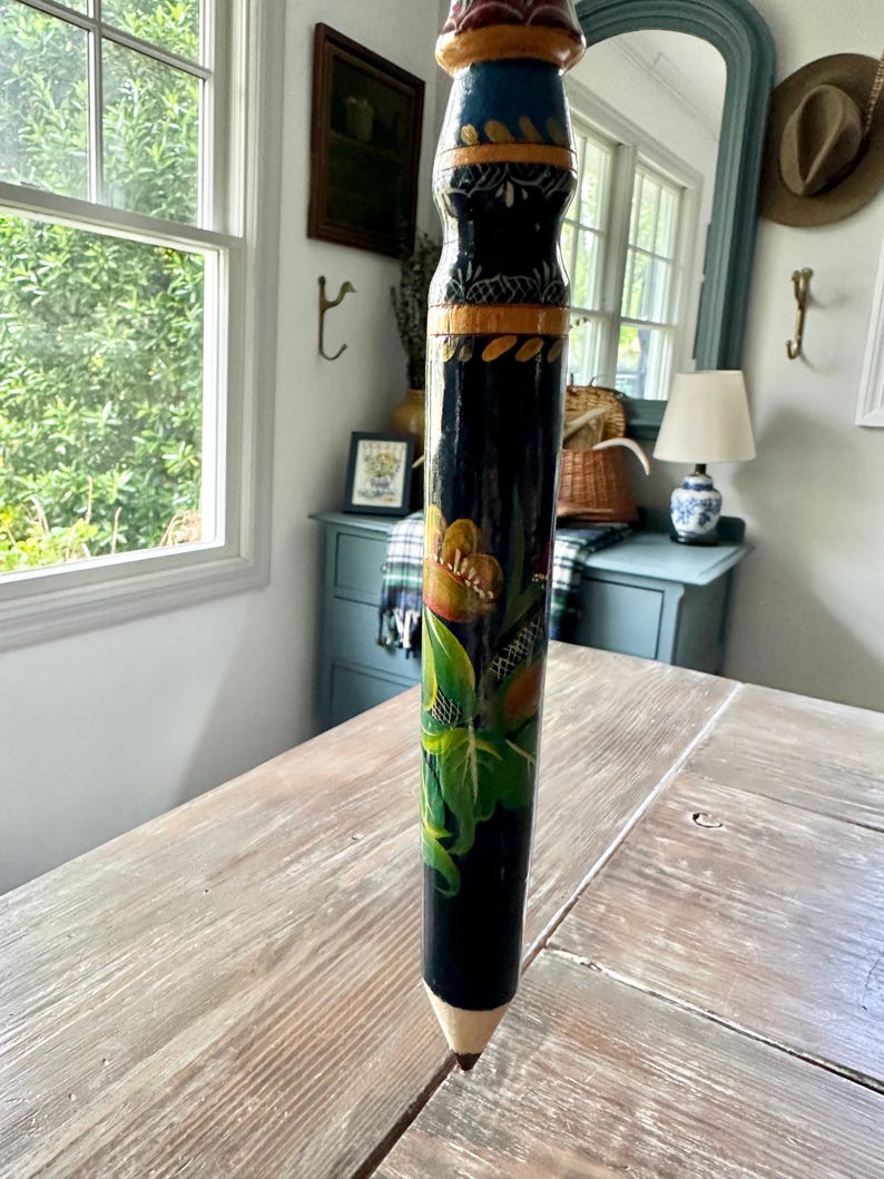 May include: A tall, decorative pencil with a painted floral design. The pencil is primarily black with colorful accents of orange, green, and blue. The pencil's tip is a natural wood color, and the top is painted with red and gold details.