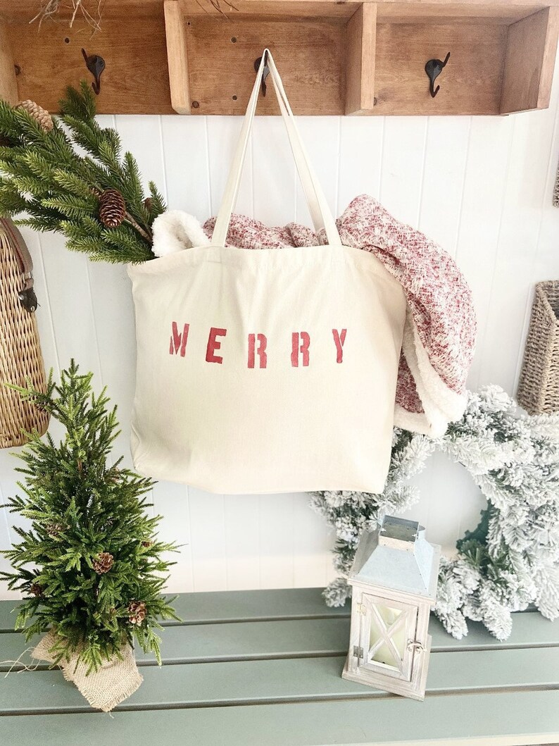May include: A white canvas tote bag with the word "MERRY" printed in red on the front. The bag is hanging on a wooden hook and has a red and white blanket inside.