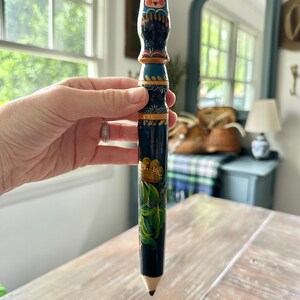 May include: A hand-painted, decorative pencil featuring a Matryoshka doll design at the top. The pencil is primarily dark blue with gold and green floral accents. The pencil has a wooden tip.