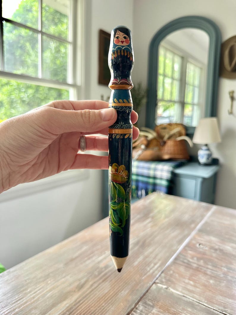 May include: A hand-painted, decorative pencil featuring a Matryoshka doll design at the top. The pencil is primarily dark blue with gold and green floral accents. The pencil has a wooden tip.