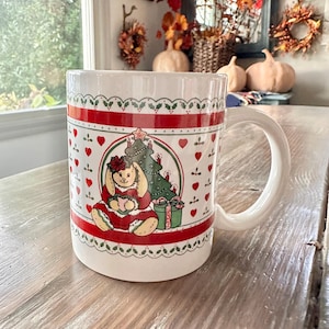 May include: White ceramic mug with a Christmas-themed design. The mug features a red and green color scheme with a bunny, Christmas tree, and gift box illustration. The mug has a white handle and a decorative border.