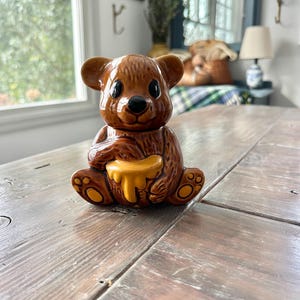 May include: A brown ceramic bear-shaped honey pot with a glossy finish. The bear is holding a yellow honey dipper. The pot has black eyes and a small, round nose. The bear is sitting on a wooden table.