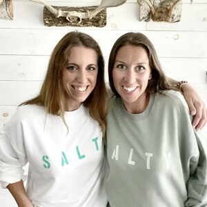 May include: Two women wearing matching sweatshirts with the word "SALTY" printed on them. The sweatshirt on the left is white with teal lettering, and the sweatshirt on the right is sage green with white lettering.