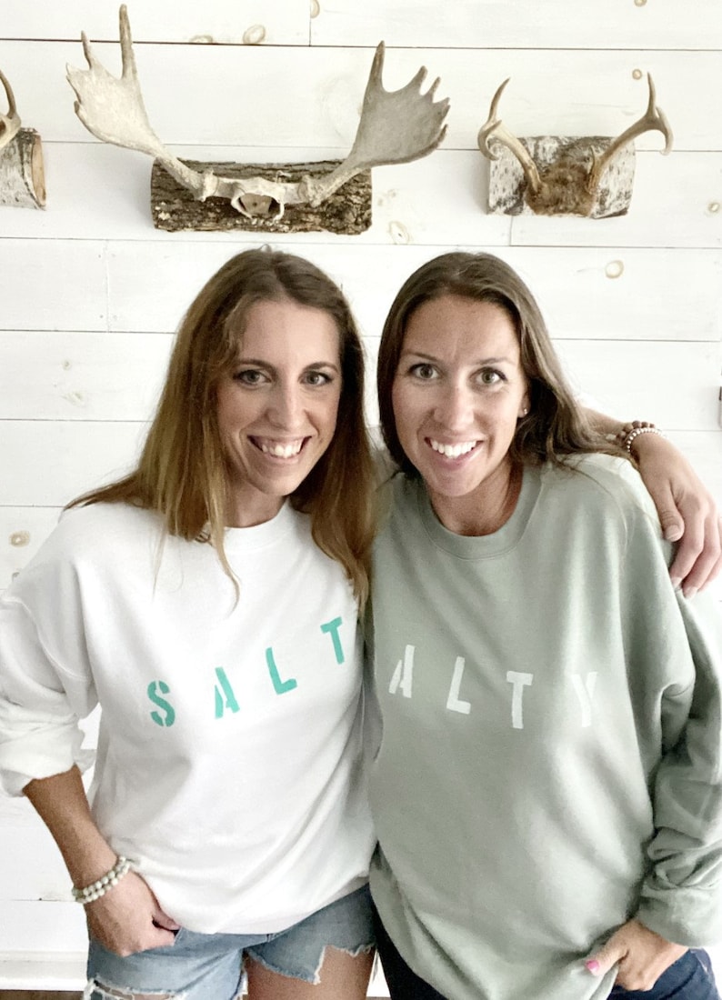 May include: Two women wearing matching sweatshirts with the word "SALTY" printed on them. The sweatshirt on the left is white with teal lettering, and the sweatshirt on the right is sage green with white lettering.