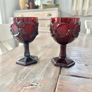 May include: Two ruby red vintage wine glasses with ornate, textured designs. The glasses have a hexagonal base and a stem that leads to a round bowl. The glasses are displayed on a rustic wooden table.