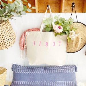 May include: A white canvas tote bag with the number 11937 printed in pink on the front. The bag is hanging on a wooden shelf with a blue and white patterned pillow in front of it.