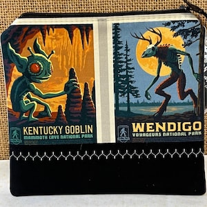 May include: A black zippered pouch with a white background featuring two colorful illustrations of mythical creatures. The left side shows a green goblin-like creature with large eyes in a cave setting. The right side shows a Wendigo, a creature with antlers and a human-like body, in a forest setting. The text "Kentucky Goblin Mammoth Cave National Park" is below the goblin illustration. The text "Wendigo Voyageurs National Park" is below the Wendigo illustration.