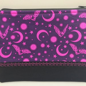 May include: A purple zippered pouch with a black bottom. The pouch has a pink and white pattern of bats, moons, and stars.
