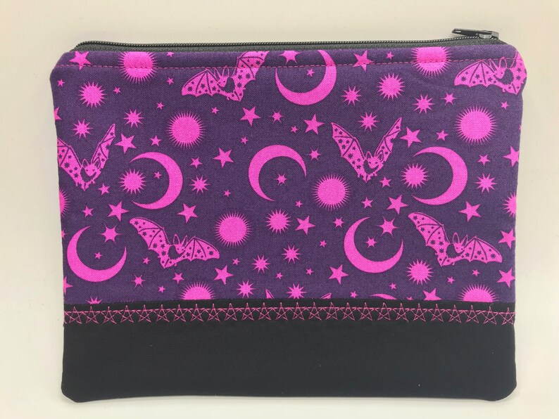 May include: A purple zippered pouch with a black bottom. The pouch has a pink and white pattern of bats, moons, and stars.