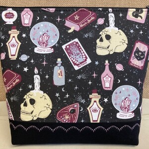 May include: Black zippered pouch with a black and white print of skulls, candles, potion bottles, tarot cards, and crystal balls. The print has a vintage, whimsical style. The pouch has a pink and black scalloped trim. The pouch has a tag that says "Avon Made".