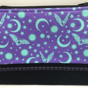 May include: Purple zippered pouch with a repeating pattern of bats, moons, and stars in a teal colour. The pouch has a black bottom with a row of silver stars.