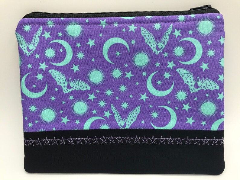 May include: Purple zippered pouch with a repeating pattern of bats, moons, and stars in a teal colour. The pouch has a black bottom with a row of silver stars.