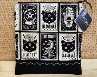 Könnte beinhalten: Schwarze und weiße Reißverschlusstasche mit einem sich wiederholenden Muster von Tarotkarten mit schwarzen Katzen, Monden und anderen mystischen Symbolen. Die Texte "The Black Cat" und "The Moon" sind auf den Karten sichtbar. Die Tasche hat einen schwarzen Hintergrund und weiße Akzente.
