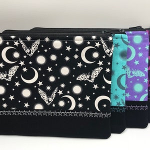 May include: Three zippered pouches with a black background and a white pattern of bats, moons, and stars. The pouches are lined up from left to right with the colours turquoise, purple, and pink.