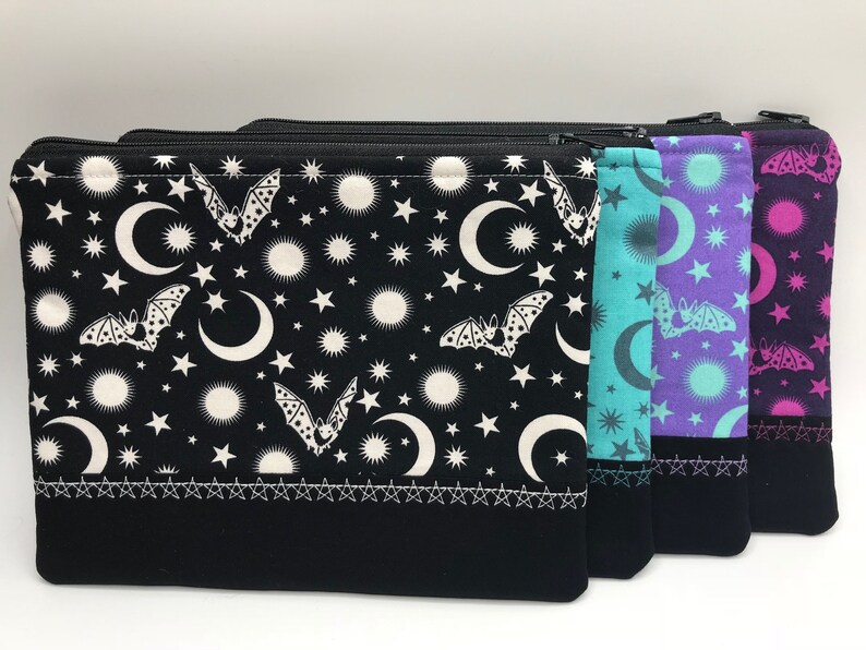 May include: Three zippered pouches with a black background and a white pattern of bats, moons, and stars. The pouches are lined up from left to right with the colours turquoise, purple, and pink.