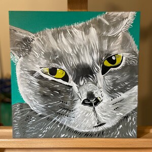 May include: Square canvas painting of a gray cat's face with yellow eyes. The cat's fur is painted in shades of gray and white, with a teal background. The painting is signed "KS 22" in the lower left corner.