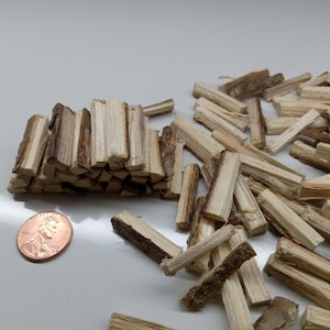 May include: A collection of small, rectangular wood pieces, some stacked and others scattered. The wood has a light brown color with darker bark-like edges. A penny is included for size reference.