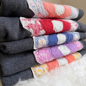 May include: A stack of five folded dark charcoal gray sweatshirts, each featuring a unique quilted panel on the front. The panels display various colors including coral, red, blue, purple, and yellow, combined with white and floral fabric.