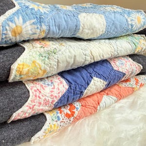May include: A stack of four folded sweatshirts, each featuring a unique quilted fabric panel on the front. The panels display various floral and geometric patterns in shades of blue, white, yellow, pink, and orange, contrasted against dark gray fabric.