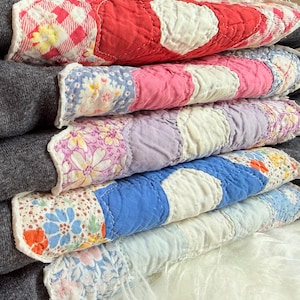 May include: A stack of folded quilts featuring alternating solid-colored and floral-patterned fabric squares. The colors include red, pink, purple, blue, and light blue, all stitched with a quilted texture and layered over dark gray fabric.