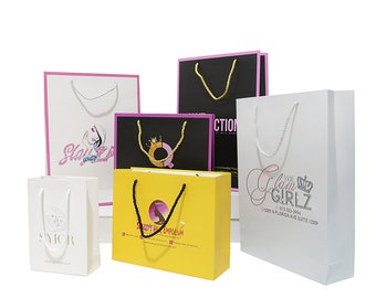 Puede incluir: Una variedad de bolsas de compras coloridas con varios logotipos y diseños. Las bolsas incluyen blanco, amarillo y negro, con detalles en rosa y dorado. Algunas bolsas presentan textos como "Glam Girlz" y "Sazzys Hair Emporium".