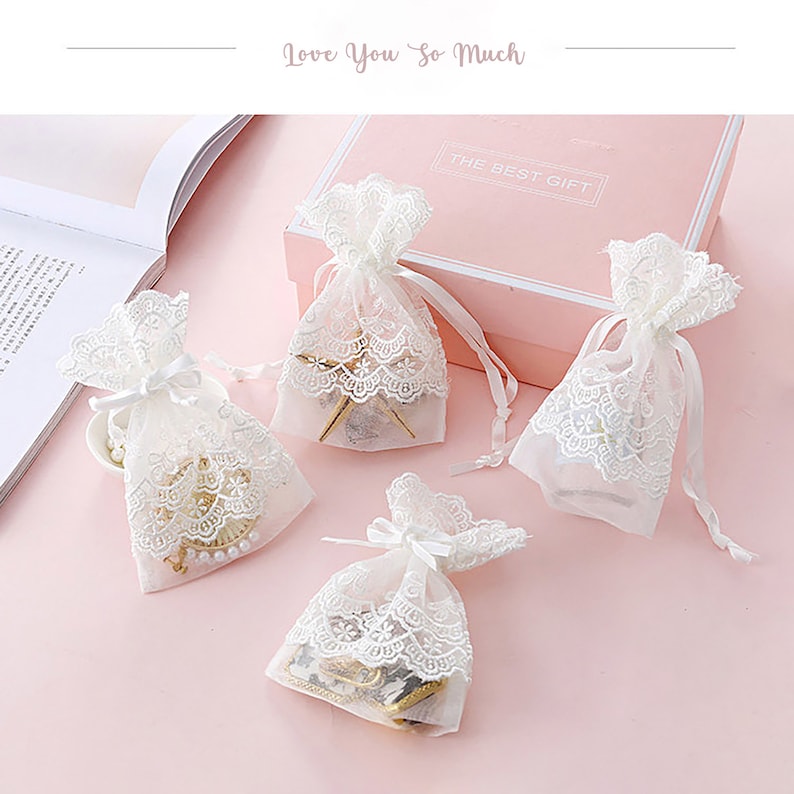 May include: Four white lace gift bags with white ribbon ties. The bags are filled with small gifts and are sitting on a pink surface. The text "The Best Gift" is visible on a white box in the background.