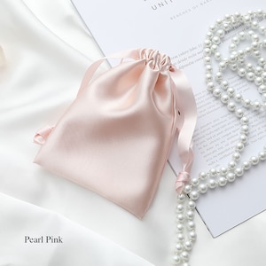 May include: A pearl pink satin drawstring bag with a white satin ribbon. The bag is lying on a white surface next to a strand of white pearls.