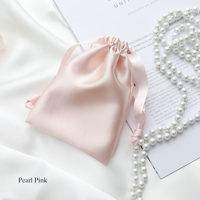 May include: A pearl pink satin drawstring bag with a white satin ribbon. The bag is lying on a white surface next to a strand of white pearls.