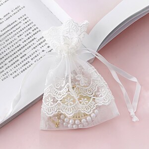 May include: A white lace drawstring bag with a pearl necklace and a gold chain inside. The bag is on a pink surface.