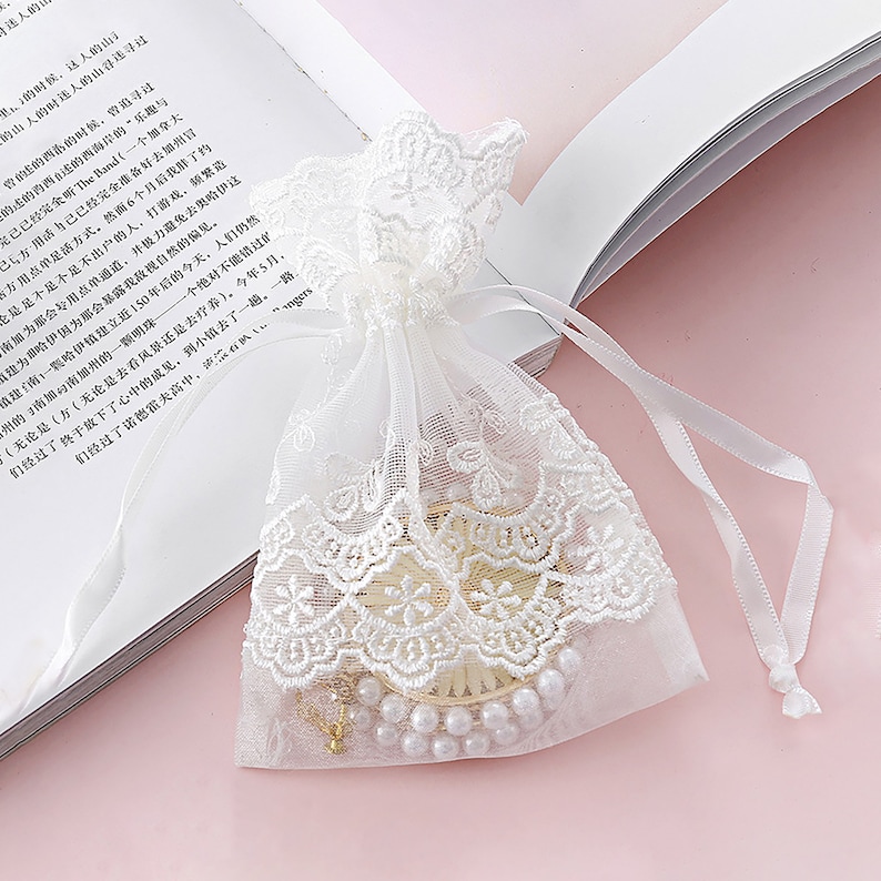 May include: A white lace drawstring bag with a pearl necklace and a gold chain inside. The bag is on a pink surface.