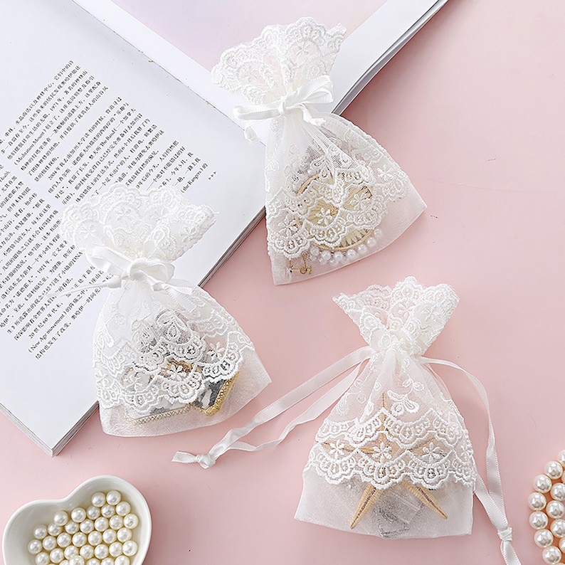 May include: Three white lace gift bags with white satin ribbon ties. The bags are filled with small decorative items. The bags are on a pink surface.