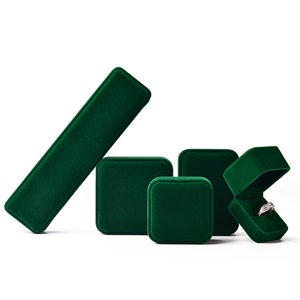 May include: A collection of emerald green velvet jewelry boxes in various shapes and sizes. One box is open, displaying a silver ring with a clear stone. The boxes are designed to hold rings, necklaces, and other jewelry items.