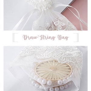 May include: White lace drawstring bag with a white ribbon. The bag is filled with white pearls and a gold-colored decorative item. The bag has a folded bottom.