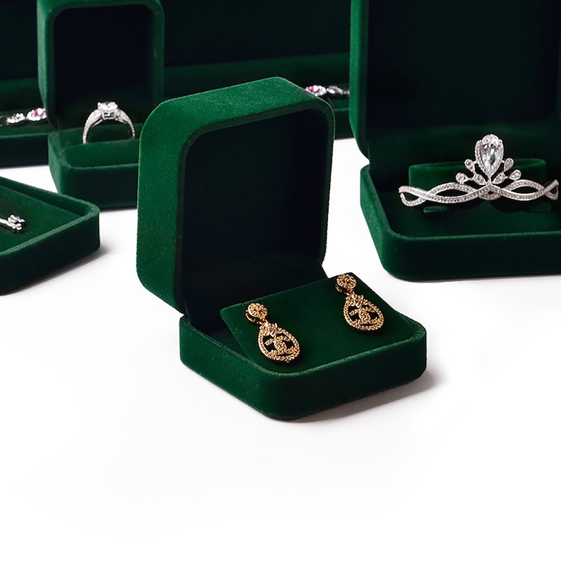 May include: Emerald green velvet jewelry boxes, some open to display rings and earrings. One box holds gold teardrop earrings with intricate detailing. Other boxes contain a diamond ring and a silver tiara. The boxes are arranged on a white surface.
