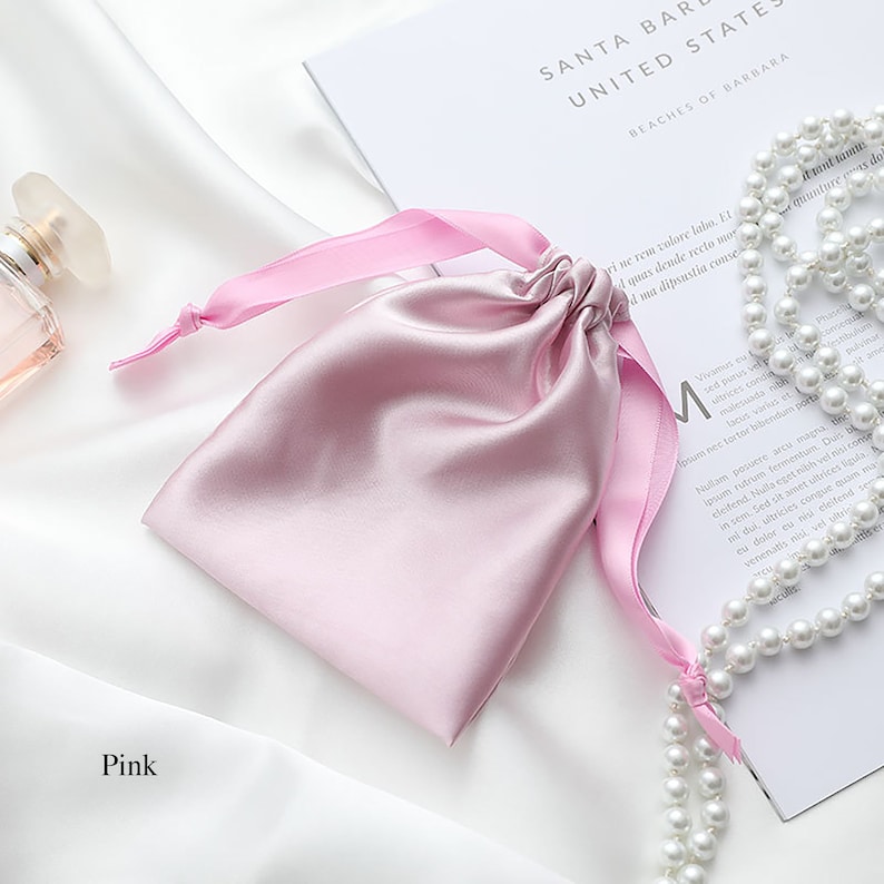 May include: A pink satin drawstring bag with a pink ribbon. The bag is on a white surface with a string of white pearls.
