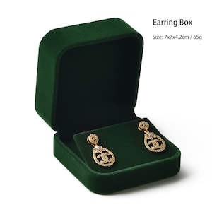 May include: A dark green velvet earring box, open to display a pair of gold-colored earrings with clear gemstones. The earrings have a teardrop shape with a decorative design. The box is approximately 7x7x4.2 cm and weighs 65g.