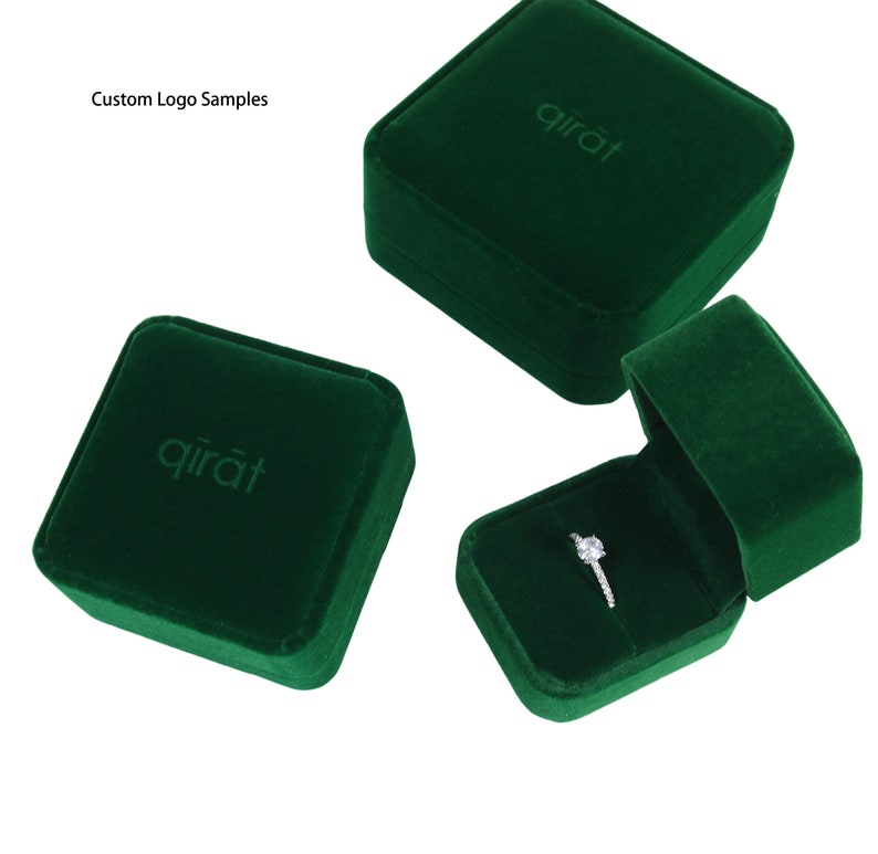 May include: Three dark green velvet ring boxes, one open to display a diamond ring. The boxes have a square shape with rounded edges and the word "qirāt" embossed on the lid. The text "Custom Logo Samples" is in the upper left corner.