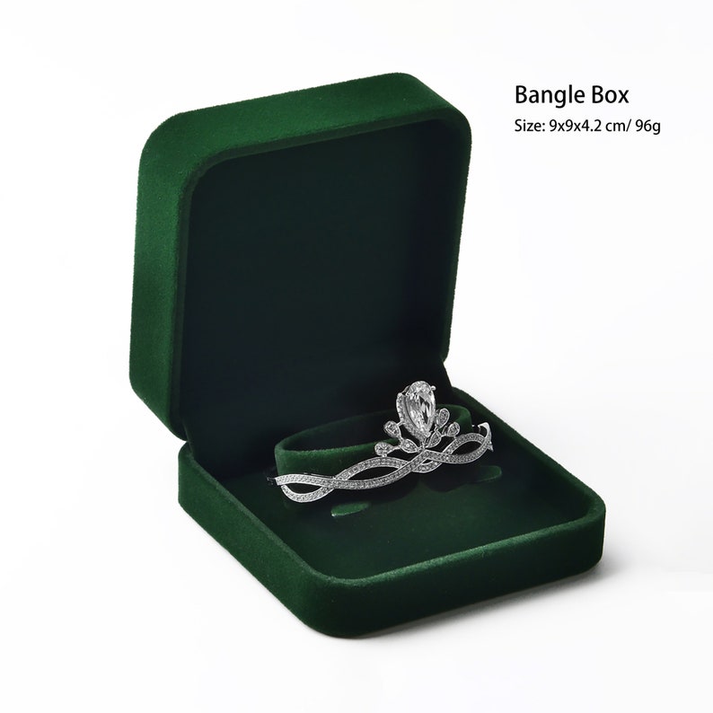 May include: A dark green velvet bangle box with a silver tiara inside. The box is open, revealing a curved holder for the tiara. The tiara is adorned with clear gemstones. The box measures 9x9x4.2 cm.