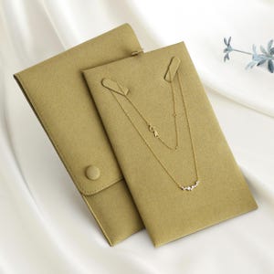 May include: A gold necklace with a delicate design is displayed on a tan jewelry card. The necklace features a series of small, sparkling stones. The card is inside a matching tan pouch with a button closure. The background is a white, silky fabric.